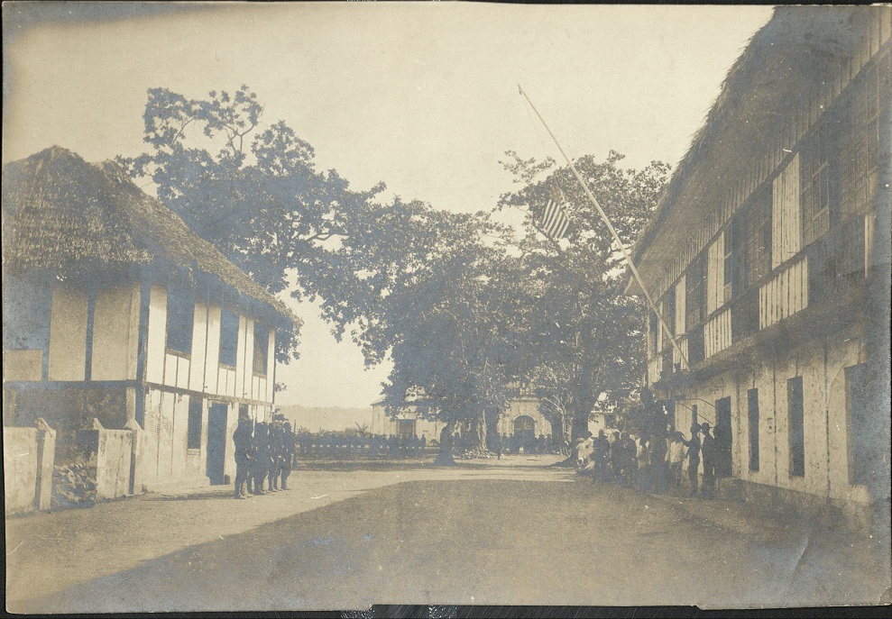Raising the flag in Cagayan. Public building on right. “L”Co. quarters on left. Church in front.