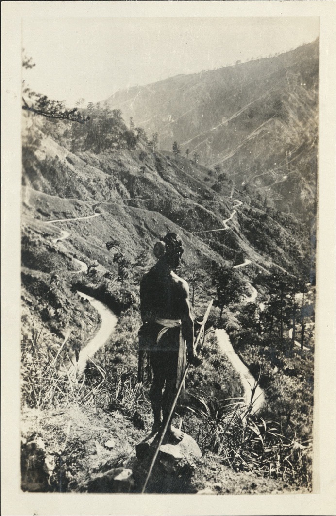 An Igorotte [Igorot] warrior in full head hunter’s regalia looking down trail on the hair pin turn of the Benguet Trail. …