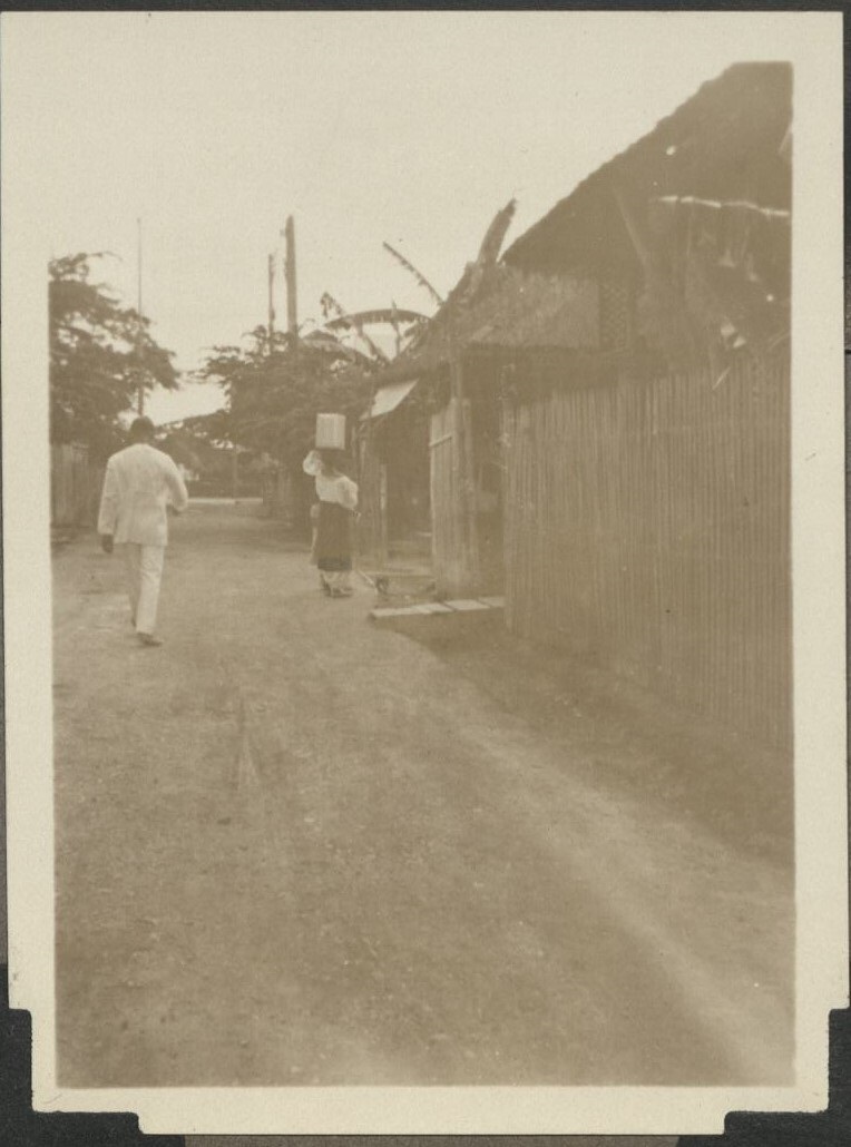 Barrio street scene, both figures in native costume. Note woman is carrying 5 gal. can of water on her head. …