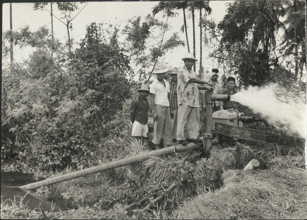 [Untitled – Men point to water flowing from a pump over a small stream].