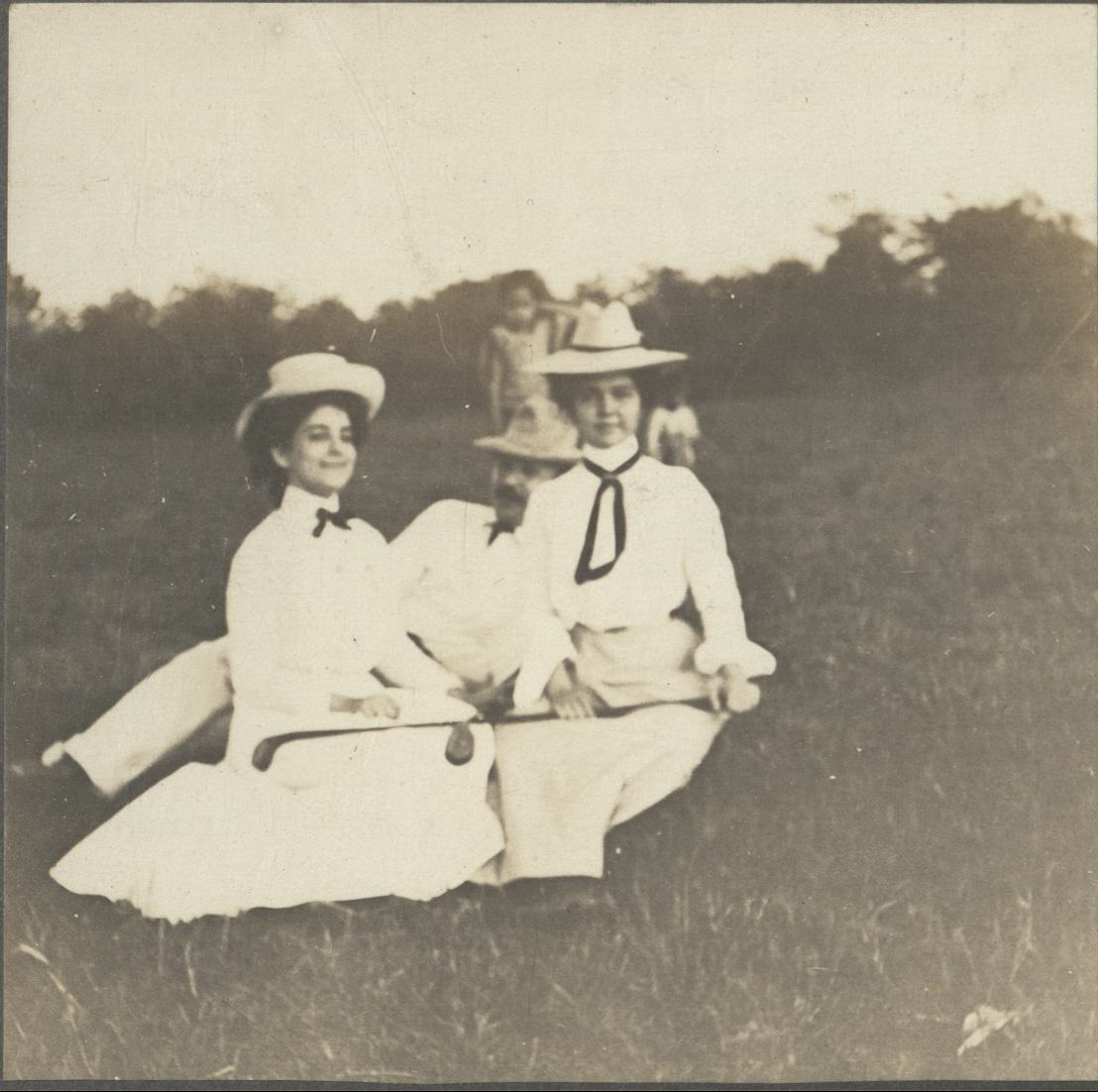 [Untitled – Two women and a man sit on grass, holding golf clubs].