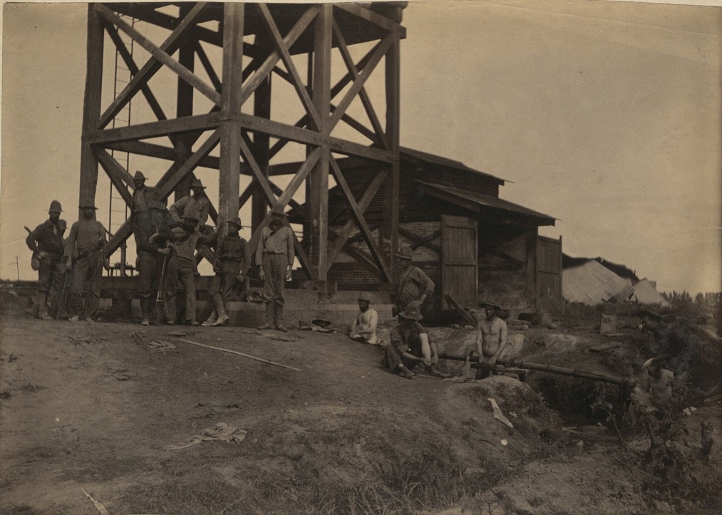 [Untitled – Soldiers stand around wooden structure and water pipe – watchtower?].