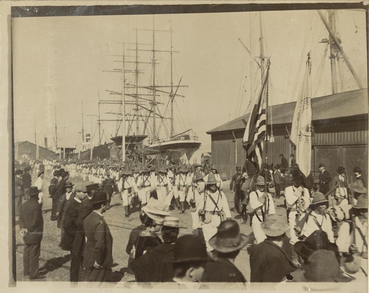[Untitled – Soldiers march in formation along a pier or wharf as civilians watch].