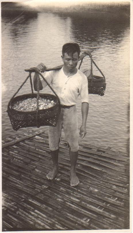 [Untitled – Man carrying baskets of small fish on a bamboo raft or dock].