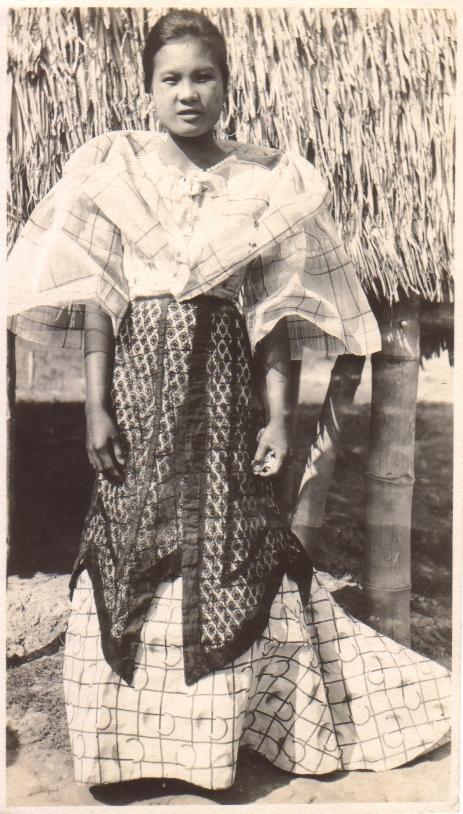 [Untitled – a Filipina in fine traditional dress poses outside a nipa hut].