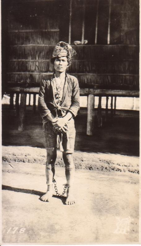 [Untitled – An indigenous man in ankle irons stands before a nipa structure].